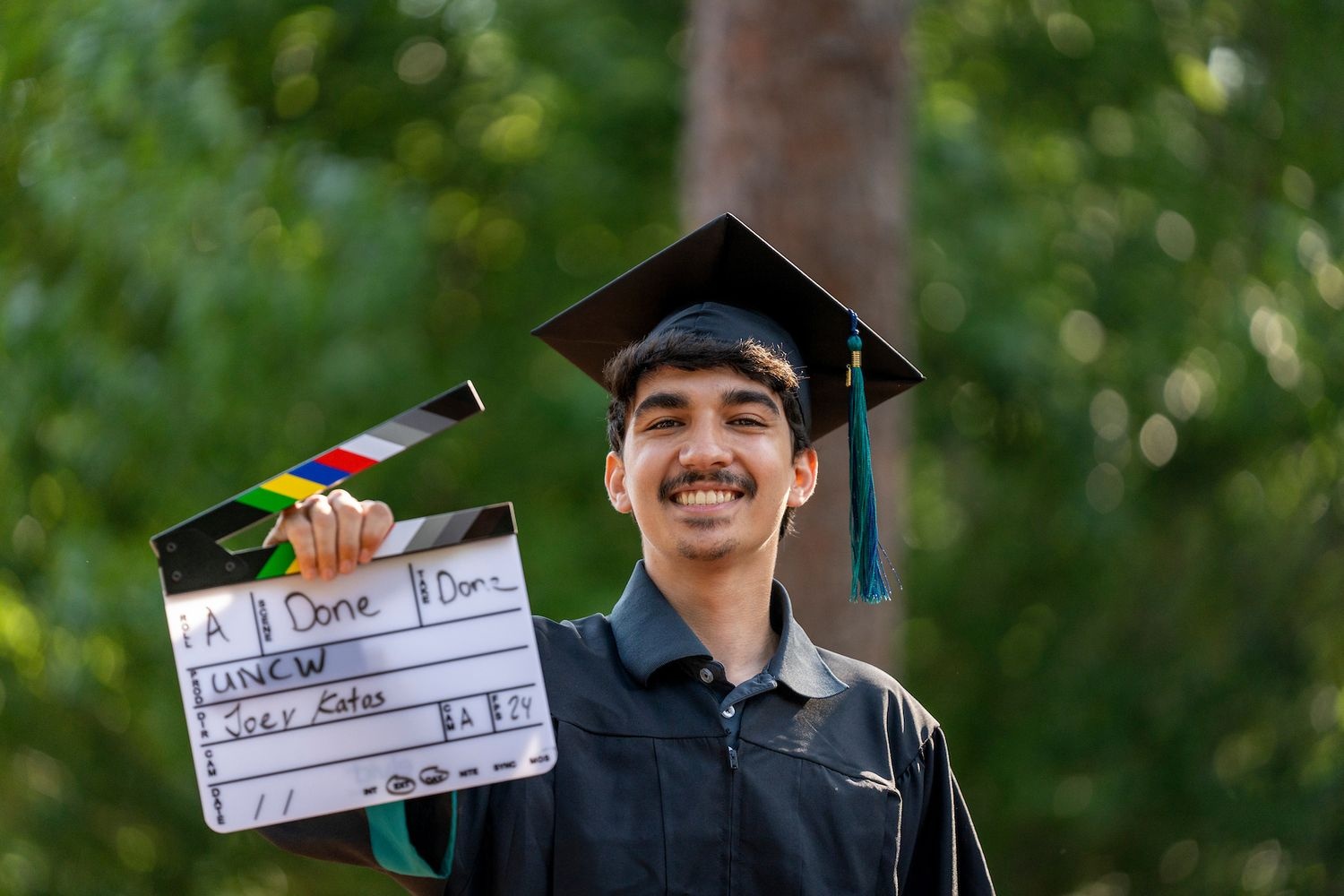 Graduating Film Studies student in Graduation regalia holding a clapperboard