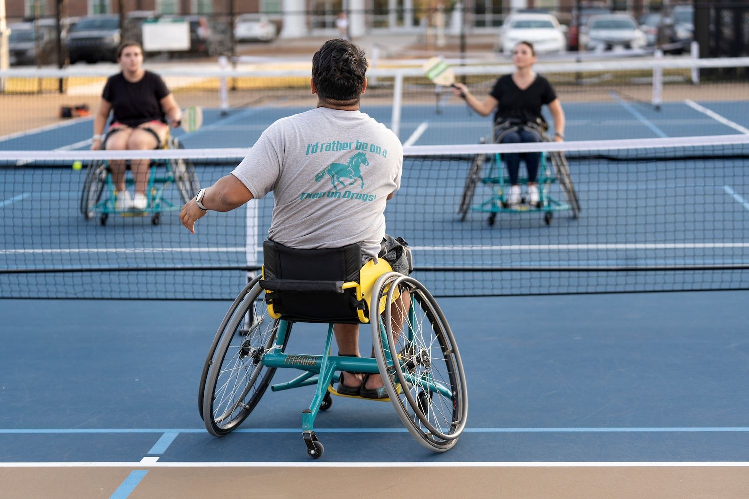One student in the foreground and two students in the background are in wheelchairs, playing pickleball on a tennis court.