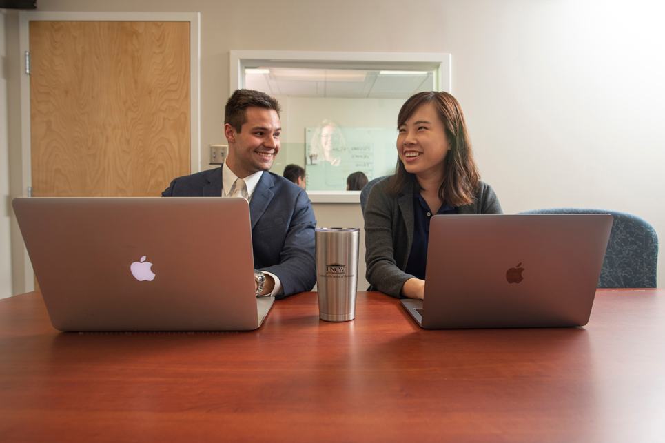 Two people are seen smiling at each other while engaging in an activity on their laptops